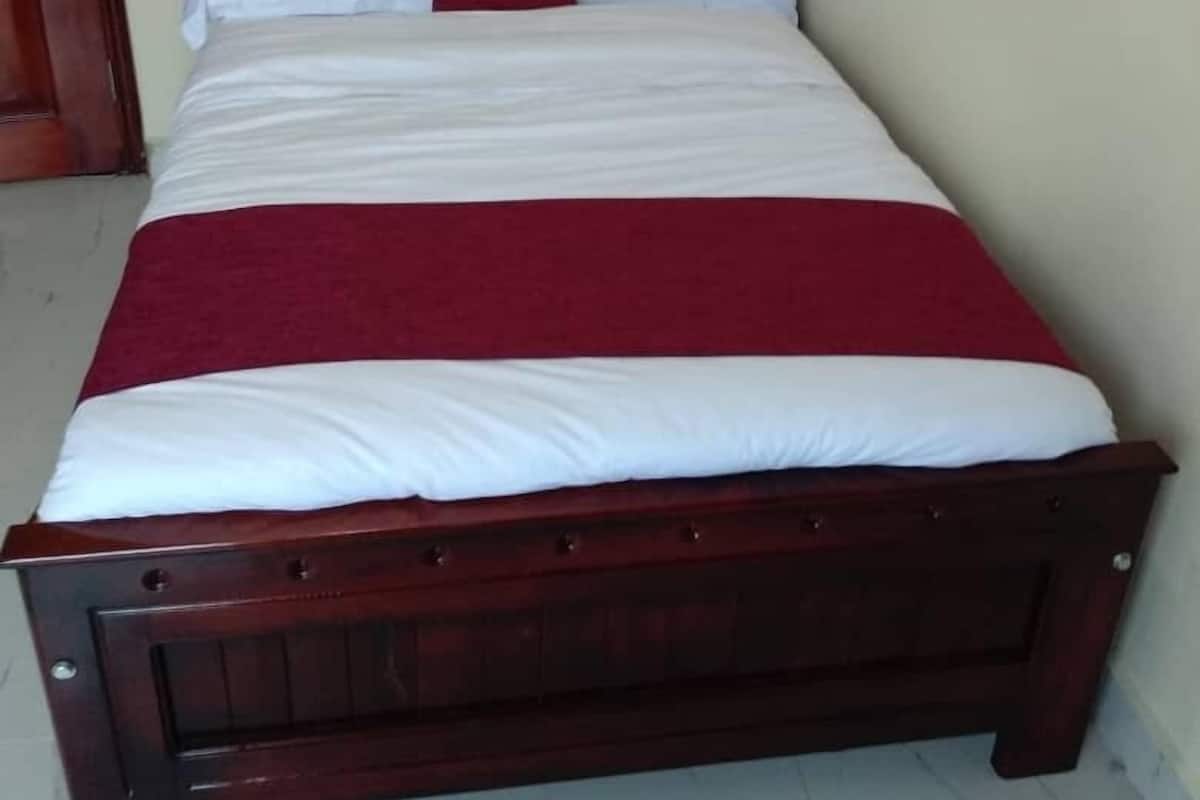 A standard single bed with a wooden frame, white and red bedding, and a plain wall with a door to the left.