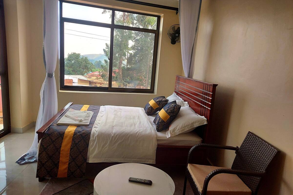 A hotel room with a bed, a chair, a round table, and a window with a view of buildings and trees.