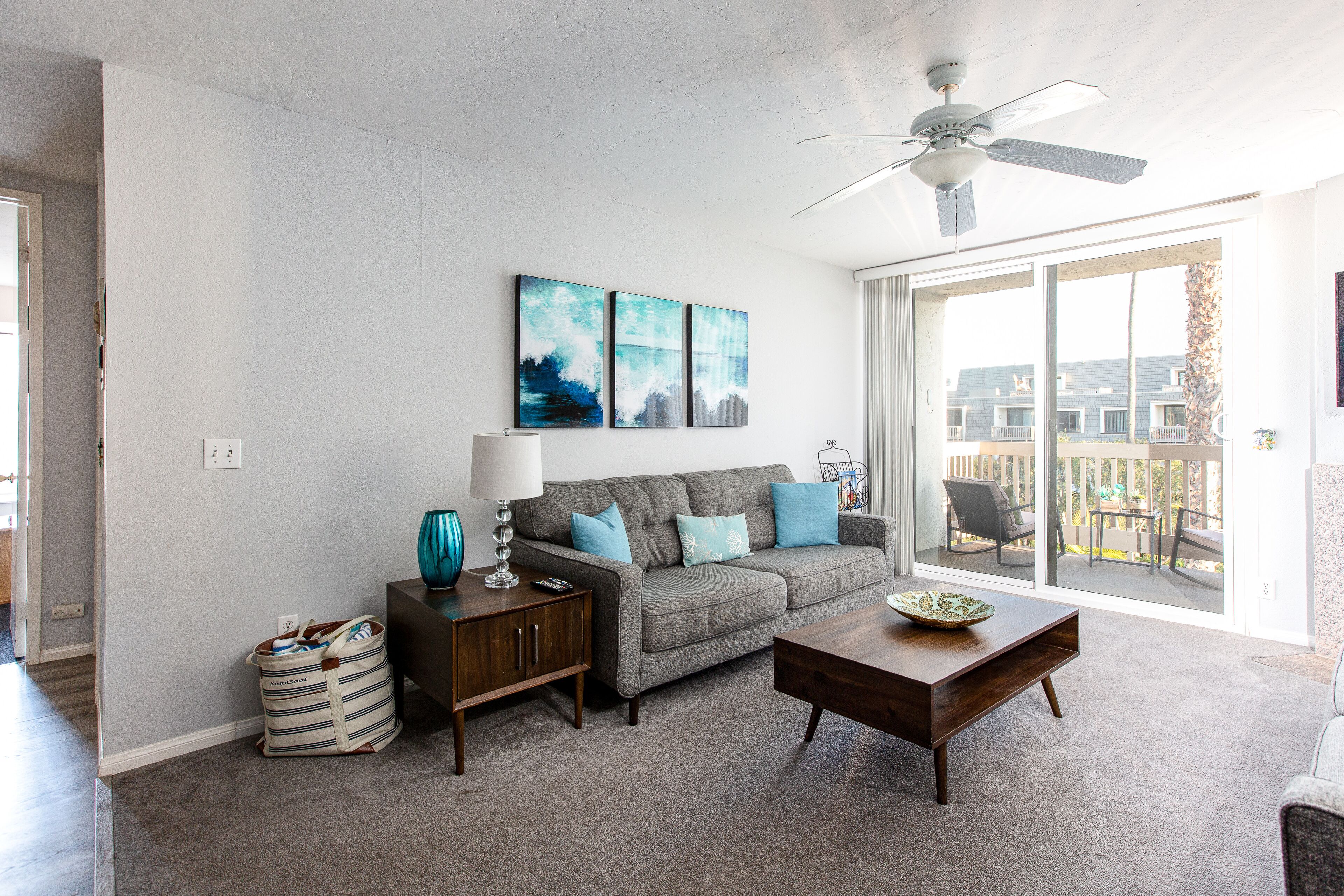 A living room with a grey sofa, a wooden coffee table, and a ceiling fan. There are two wall-mounted paintings and a bag on the floor.