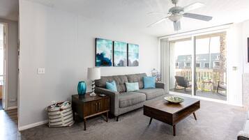 A living room with a grey sofa, a wooden coffee table, and a ceiling fan. There are two wall-mounted paintings and a bag on the floor.