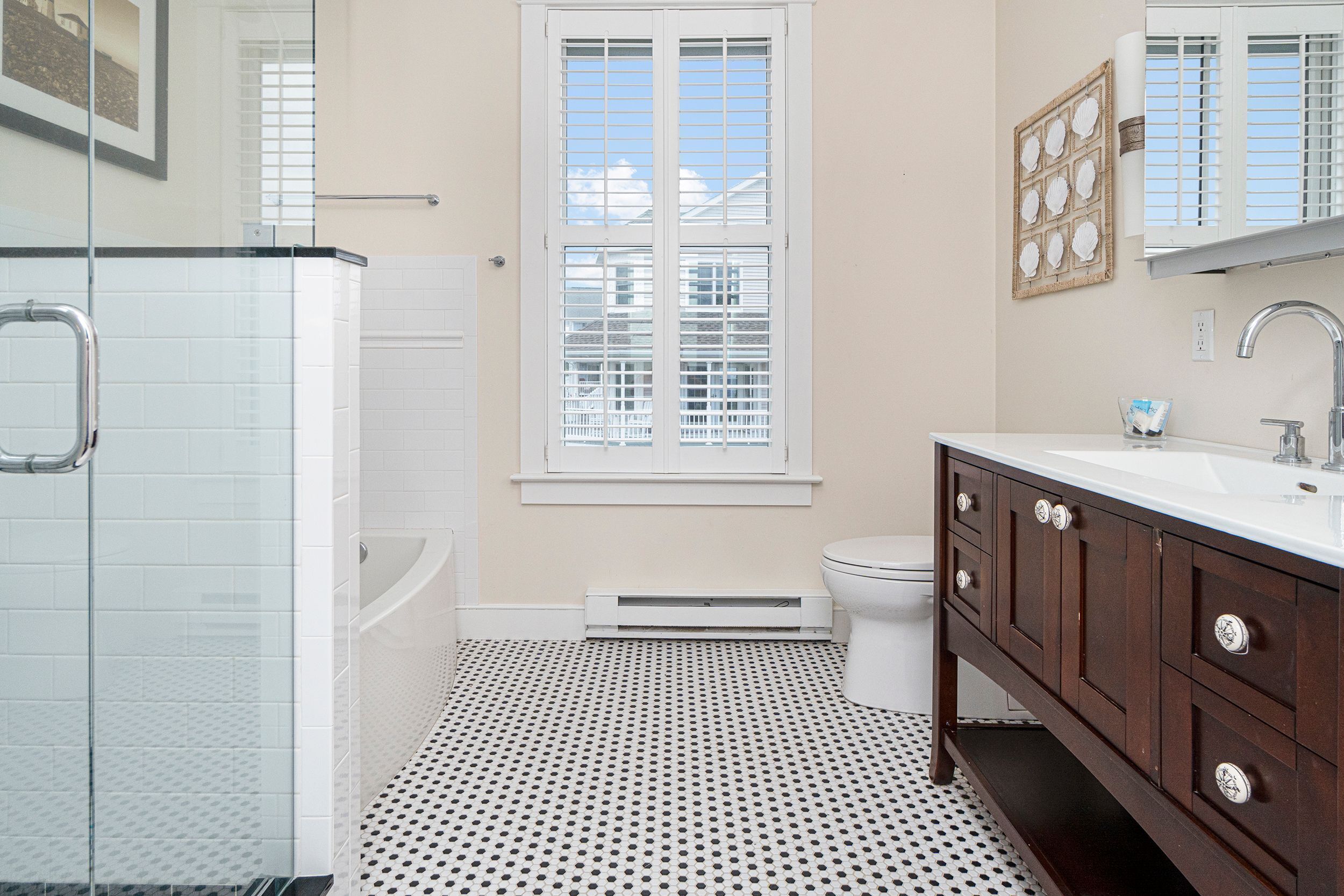 A bathroom with a glass shower enclosure, a white toilet, a white sink with a dark wooden vanity, and a window with blinds.