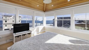 A bedroom with a large bed, a TV on a white stand, and a view of the ocean through large windows. - Panoramic Ocean View, 5 BR, Central Air, 10 minute walk to Scarborough Beach (Narragansett)