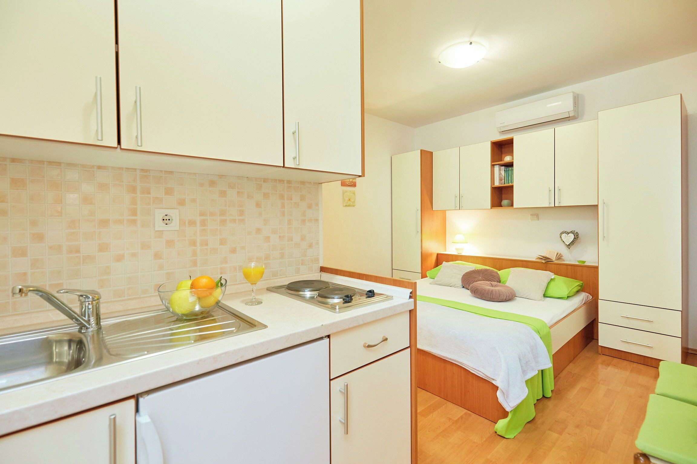 A compact kitchen with a sink, a bowl of fruit, and a glass of juice. Adjacent is a bedroom with a bed, pillows, and a nightstand.