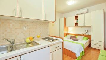 A compact kitchen with a sink, a bowl of fruit, and a glass of juice. Adjacent is a bedroom with a bed, pillows, and a nightstand.