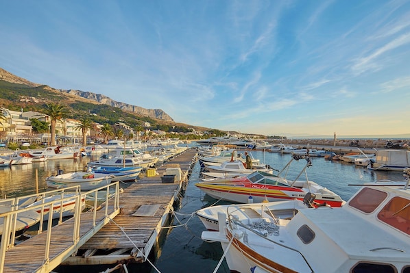 A marina with various boats docked at wooden piers, surrounded by buildings and hills in the background.