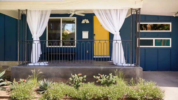 Una casa con porche cubierto, cortinas blancas y puerta amarilla.