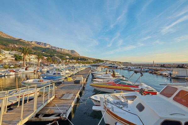 A marina with various boats docked at wooden piers, surrounded by buildings and hills in the background.