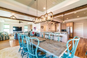 A spacious dining area with a wooden table and blue chairs, a kitchen with modern appliances, and a living room with a large TV. - Creekfront Home with Pool, Dock and Stunning Views (Pawleys Island)
