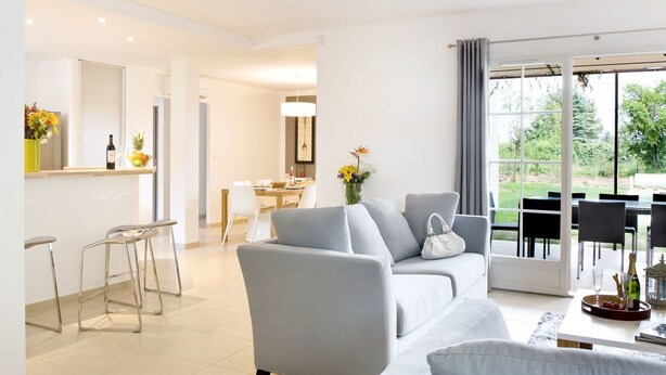 Ein modernes Wohnzimmer mit grauem Sofa, einem Essbereich mit Tisch und Stühlen sowie einer Küchenzeile mit Theke und Hockern.