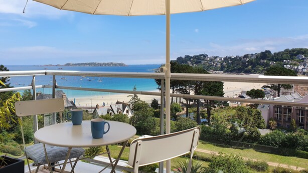 Uma varanda com mesa e cadeiras, com vista para uma praia e uma cidade costeira.