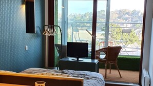 Quarto de hotel moderno com área de jantar, cama, varanda com vista para a cidade e uma janela grande.