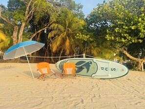 A beach with two orange beach chairs, a blue and white beach umbrella, and a surfboard leaning against a white structure. - Anantasun | Signature Pool View King Suite (Roatán)