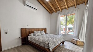 Een slaapkamer met een houten bed, een raam met gordijnen, een klein nachtkastje en een wandairconditioner.