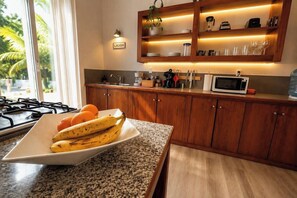 A kitchen with wooden cabinets, a granite countertop, a microwave, and a plate of bananas and oranges. - Anantasun | Signature Pool View King Suite (Roatán)