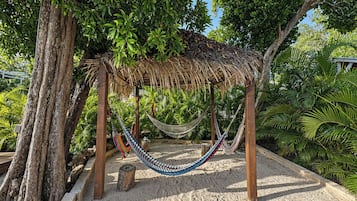 A hammock under a thatched structure surrounded by trees and sand.