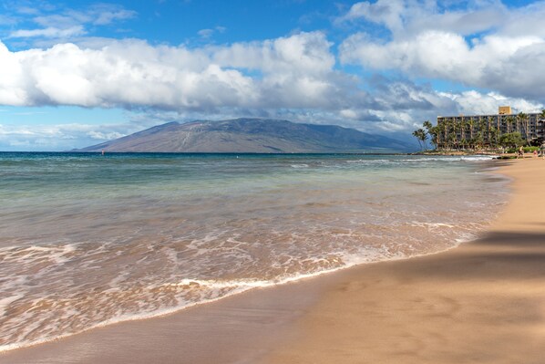 Una playa con aguas cristalinas, una montaña al fondo y un hotel con piscina y palmeras.