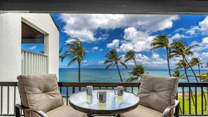 Ein Balkon mit zwei Korbstühlen und einem Glastisch, der auf einen Strand mit Palmen und das Meer blickt.