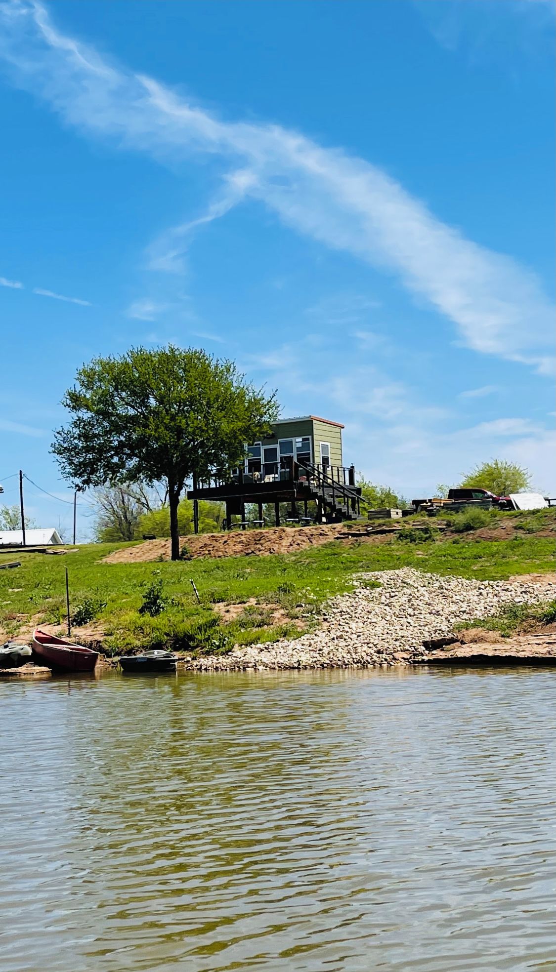 A house on a grassy hill with a tree in front, overlooking a body of water.