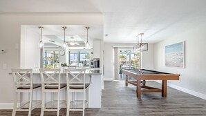 A modern kitchen with white cabinetry, a bar area with white chairs, and a pool table.