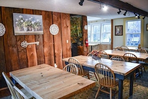 A rustic dining area with wooden tables and chairs, a painting of flowers, and a forest mural on the wall. - Lavender / Brook Road Inn (Goshen)