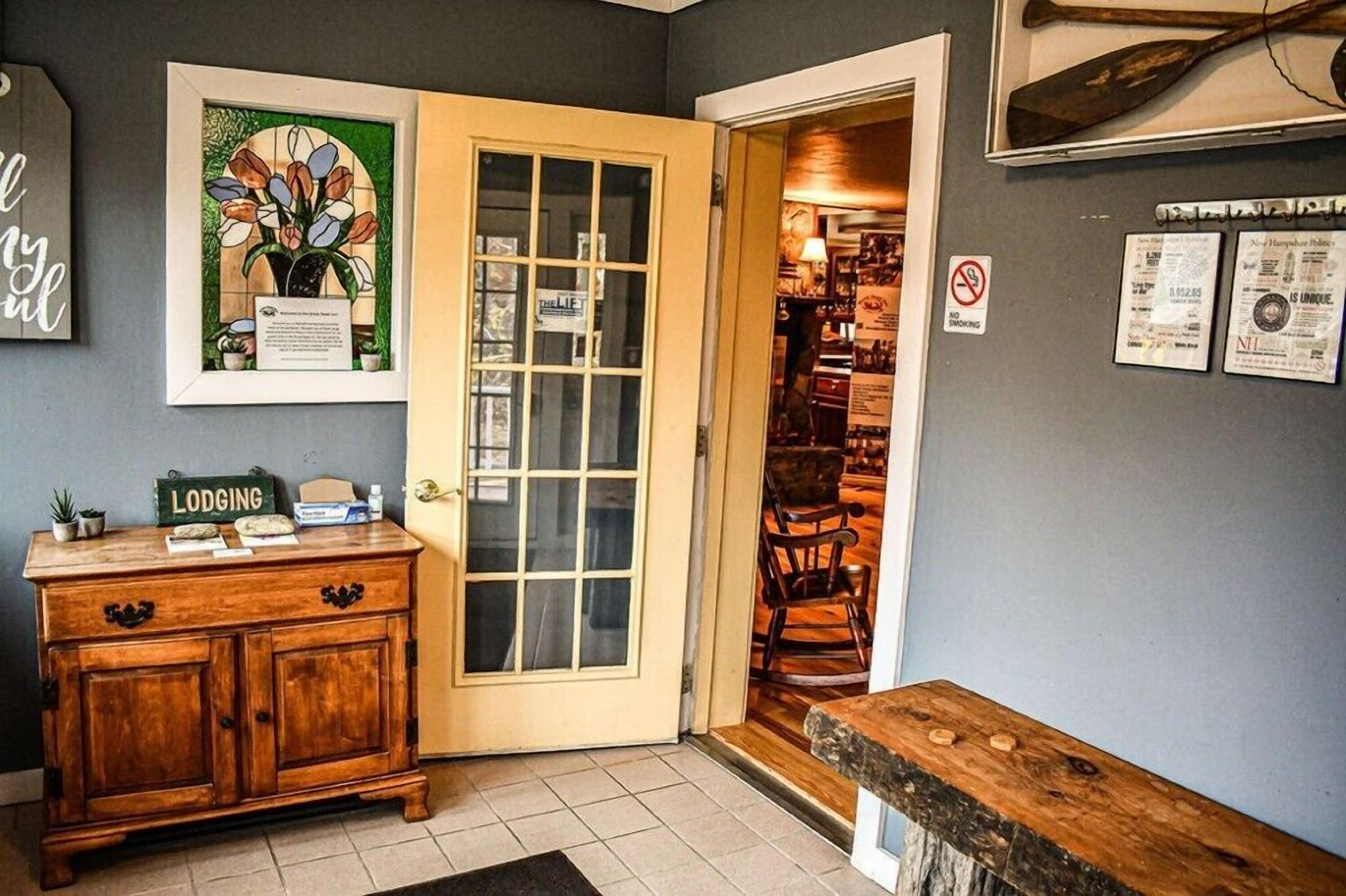 A wooden cabinet with a 'Lodging' sign, a framed floral artwork, and a door leading to a room with a chair and a wooden bench.