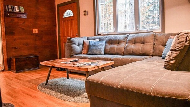 A cozy living room with a large sectional sofa, a wooden coffee table, and a view of trees through a window. - Brook Road Cabin (Goshen)
