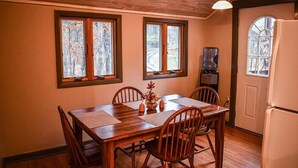 A dining area with a wooden table and chairs, two windows with a view of trees, a water dispenser, and a refrigerator. - Brook Road Cabin (Goshen)