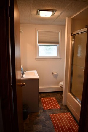 A bathroom with a white sink, toilet, and shower. There is a window above the toilet and a door on the left. - Brook Road Condo (Goshen)