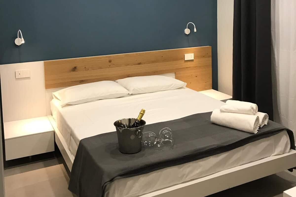 A modern hotel room with a wooden headboard, a bed with white linens, a bedside table with a lamp, and a nightstand.