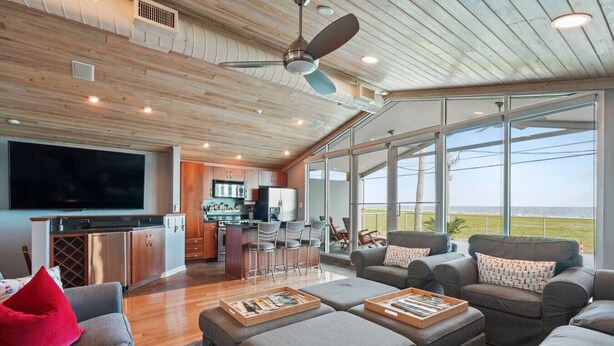 A spacious living area with a large TV, comfortable seating, a kitchen, and large windows offering a view of the outdoors.