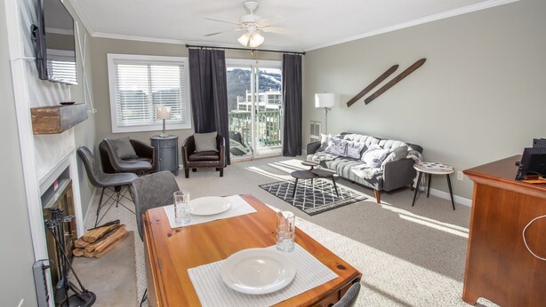 A modern living room with a sofa, chairs, a dining table set for two, a ceiling fan, and a view of the outdoors. - Ski Condo on Beech Mountain (Beech Mountain)