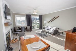 A modern living room with a sofa, chairs, a dining table set for two, a ceiling fan, and a view of the outdoors.