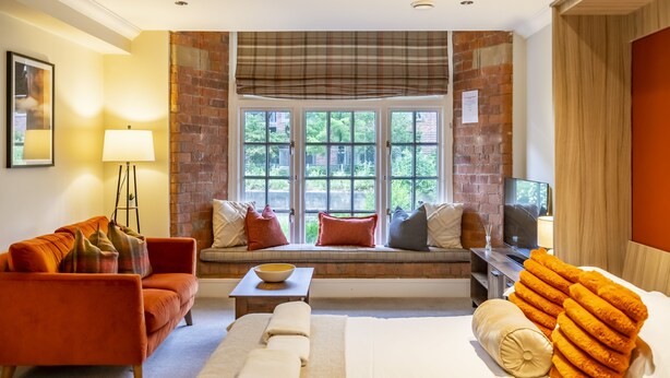 A hotel room with a sofa, a bed, a window seat with a television, and a view of greenery outside.