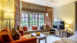 A hotel room with a brick wall, large window, sofa, TV, bedside table, and lamp.