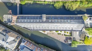 An aerial view of a long, rectangular building with a flat roof, adjacent to a river, with parking spaces and greenery around.