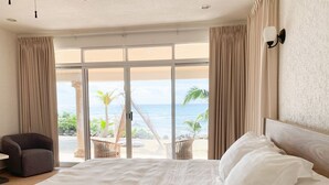 Ein Hotelzimmer mit einem großen Bett, einem Sessel und Blick auf den Strand und das Meer durch eine Schiebetür.