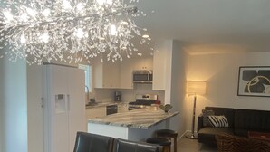 Eine moderne Küche mit weißen Schränken, einer zentralen Kochinsel und einer Kronleuchter. Es gibt einen Essbereich mit Tisch und Stühlen sowie ein Wohnzimmer mit Sofa und Lampe.