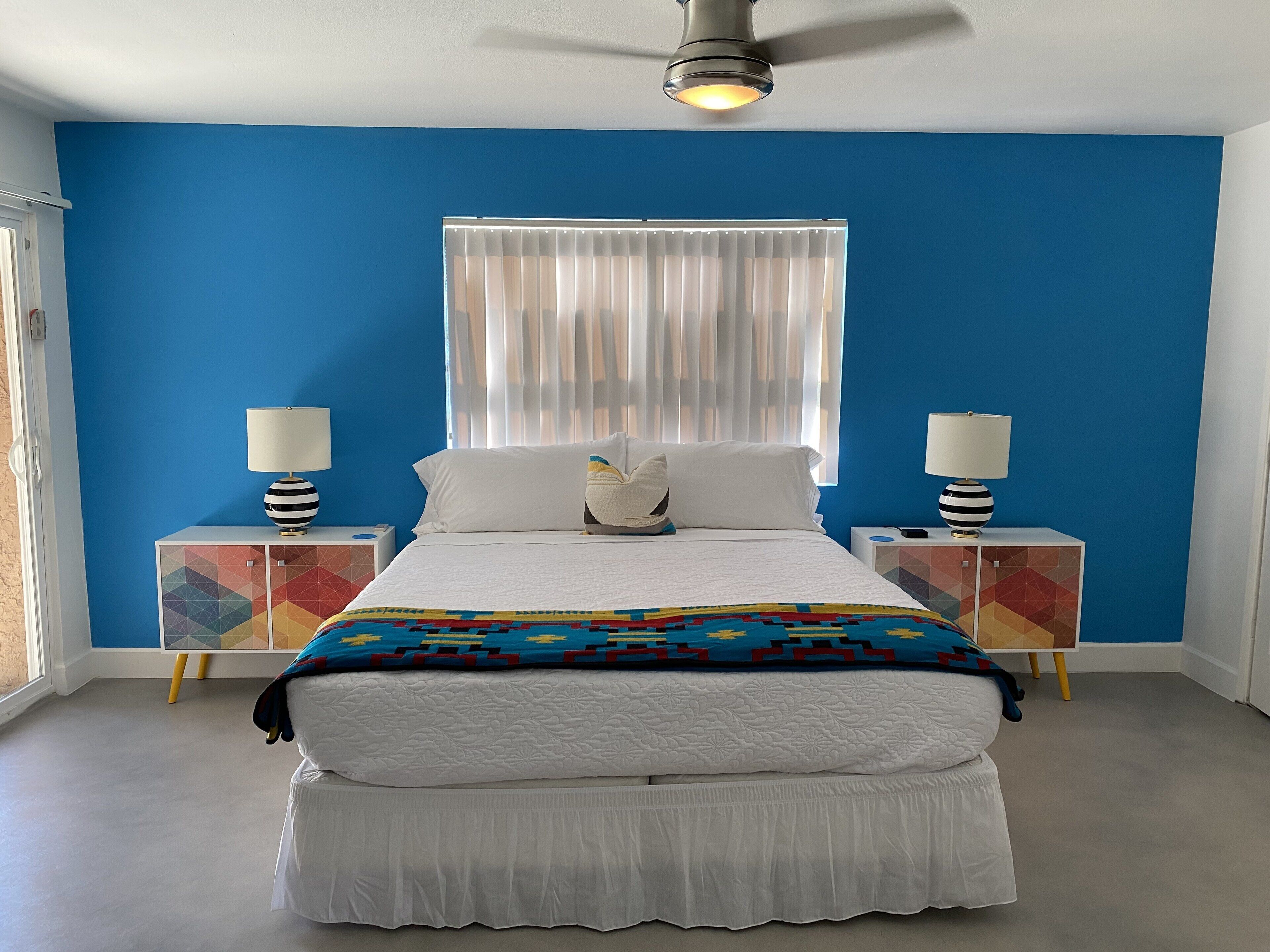 A bedroom with a bed, two bedside tables, a ceiling fan, a window with curtains, and a colorful geometric-patterned cabinet.