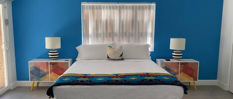 A bedroom with a bed, two bedside tables, a ceiling fan, a window with curtains, and a colorful geometric-patterned cabinet.