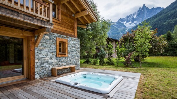 A wooden cabin with a stone wall and a hot tub on a wooden deck, surrounded by a lush garden and mountains in the background. - Chalet Rass: Sublime five bedroom chalet fully equipped (Chamonix)