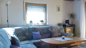 A living room with a sofa, a table, a lamp, a TV, and a window with plants.