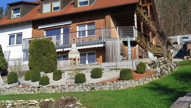 A two-story wooden house with a red-tiled roof, a stone retaining wall, and a small garden with trimmed bushes.