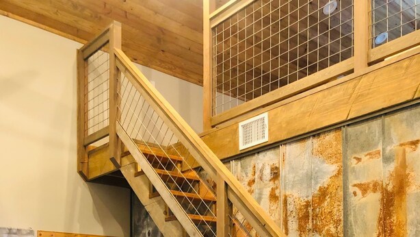 A staircase with wooden railings and a metal grid, leading to a room with a rustic wall and a dining area with a table and chairs. - Gone West Ranch cabin in Fredericksburg  (Fredericksburg)