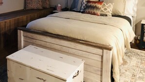 A bedroom with a wooden ceiling, a bed with multiple pillows, a bedside table, and a small white chest of drawers. - Gone West Ranch cabin in Fredericksburg  (Fredericksburg)