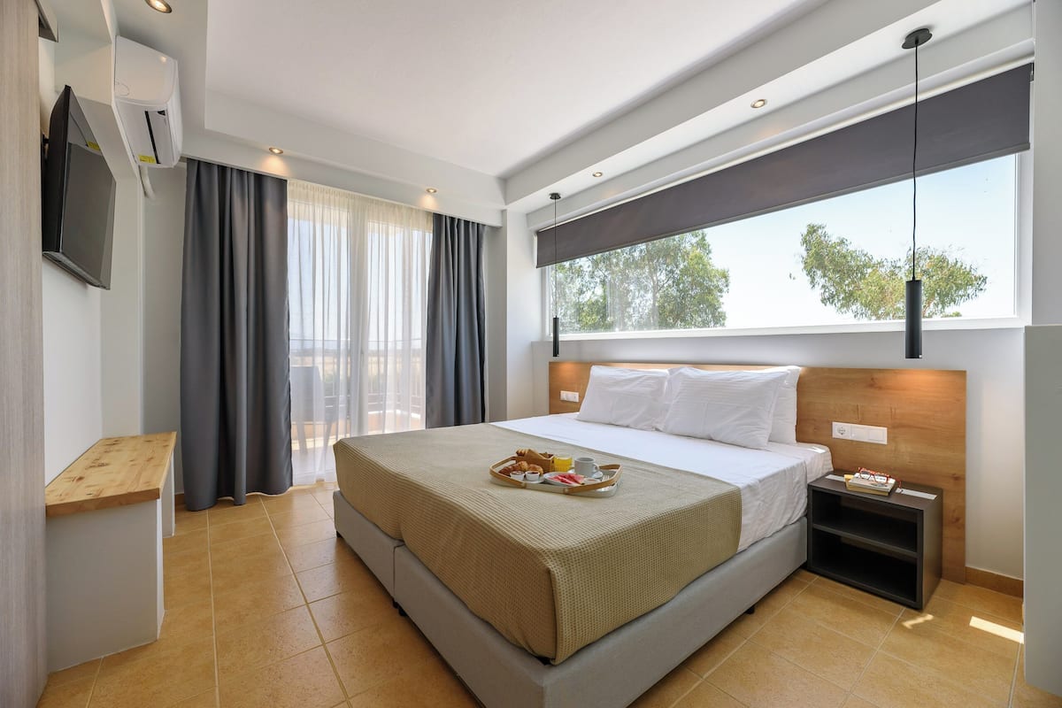 A modern hotel room with a large bed, a bedside table, a TV, and a window with a view of trees.