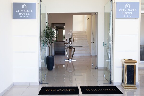 A hotel entrance with a "Welcome" mat, a potted plant, a reception desk, and a staircase leading to the upper floors. - City Gate Hotel Airport Thessaloniki (Thermi)