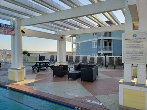 A covered pool area with lounge chairs and tables, a pool sign, and a lifeguard warning sign.