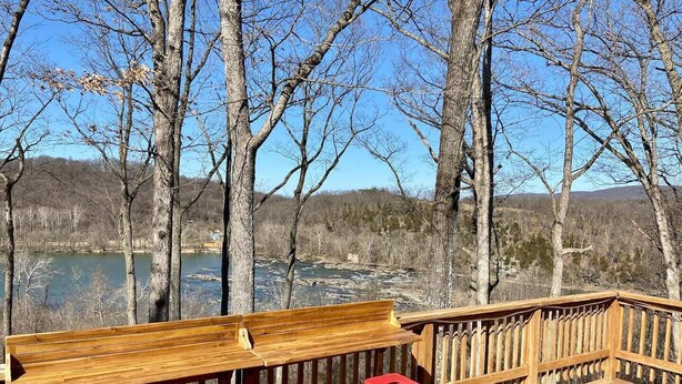 Una terraza de madera con un banco y taburetes rojos, con vistas a un lago rodeado de árboles.