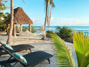 Une plage avec un abri de chaume, un hamac et des chaises longues dotées de coussins.