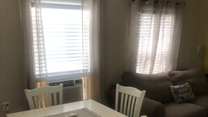 Zona de menjador amb taula i cadires blanques, un sofà marró amb coixí, i una finestra amb cortines.
